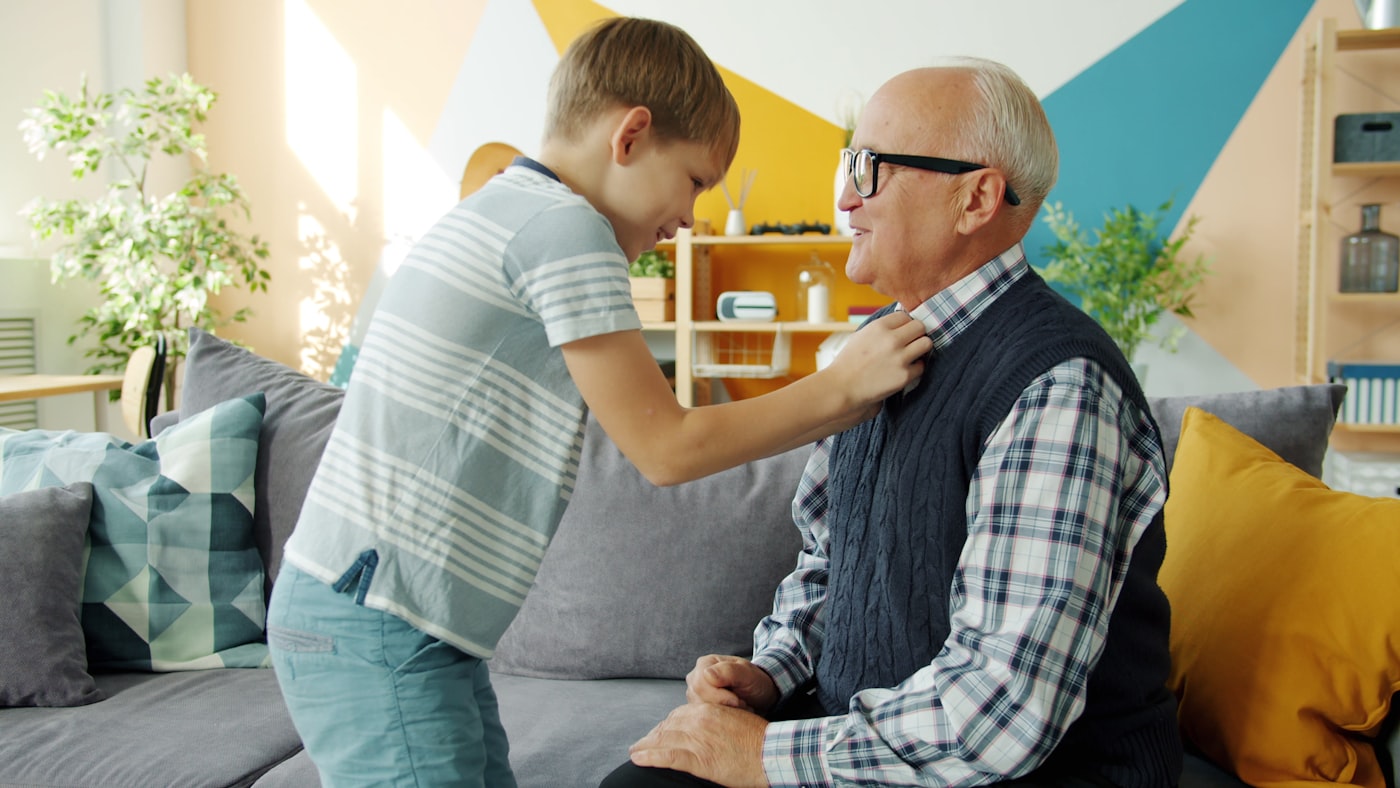 A young boy gently adjusting his grandfather's collar while the grandfather smiles, at home on a sofa.