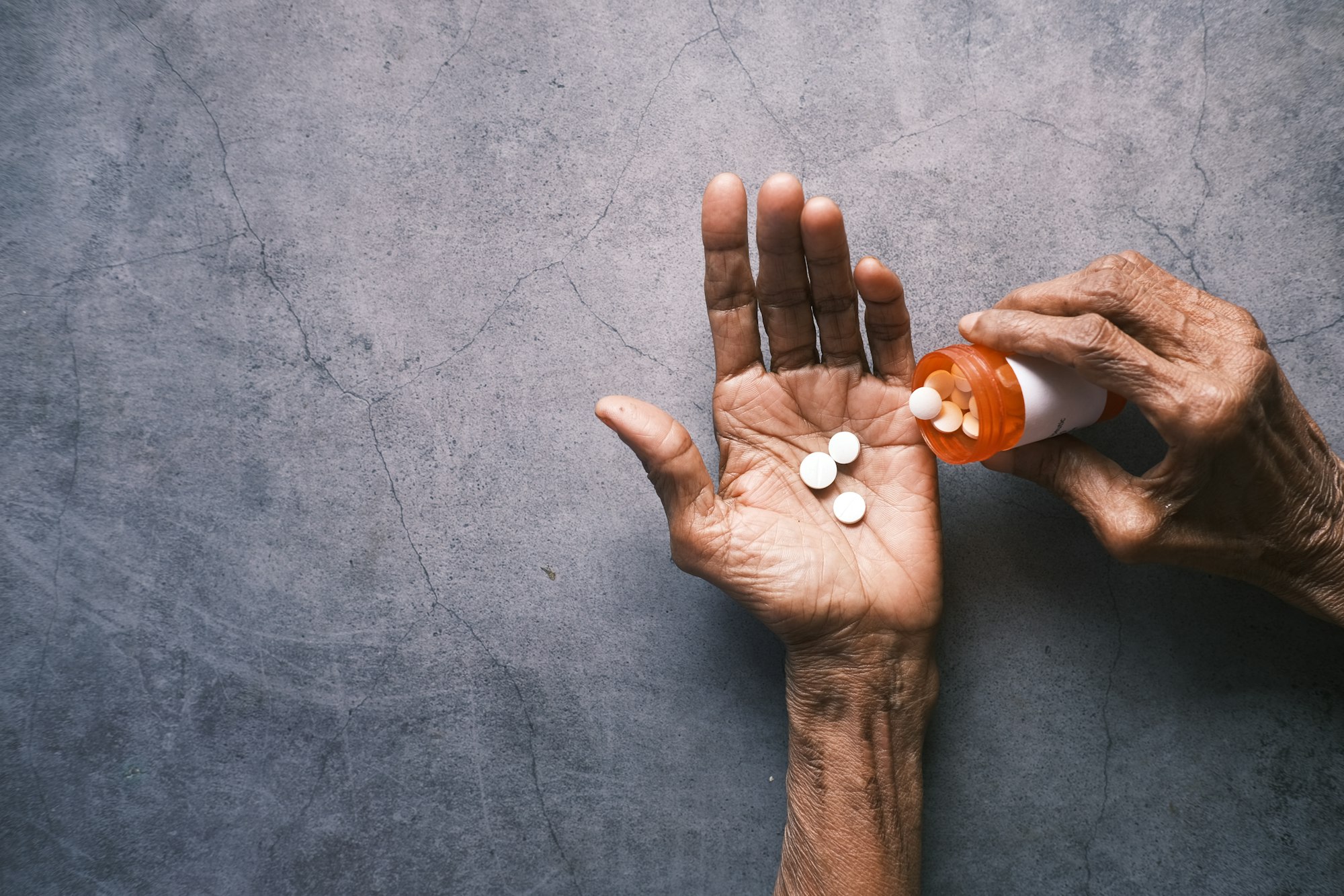 An older person's hands tipping white tablets from an orange bottle into an open palm, on a slate surface.