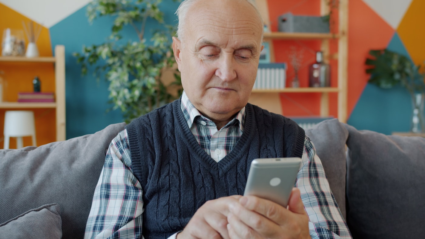 An older man reading his phone on a sofa at home.